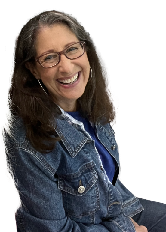 A woman wearing glasses and jeans is smiling at the camera while indoors.